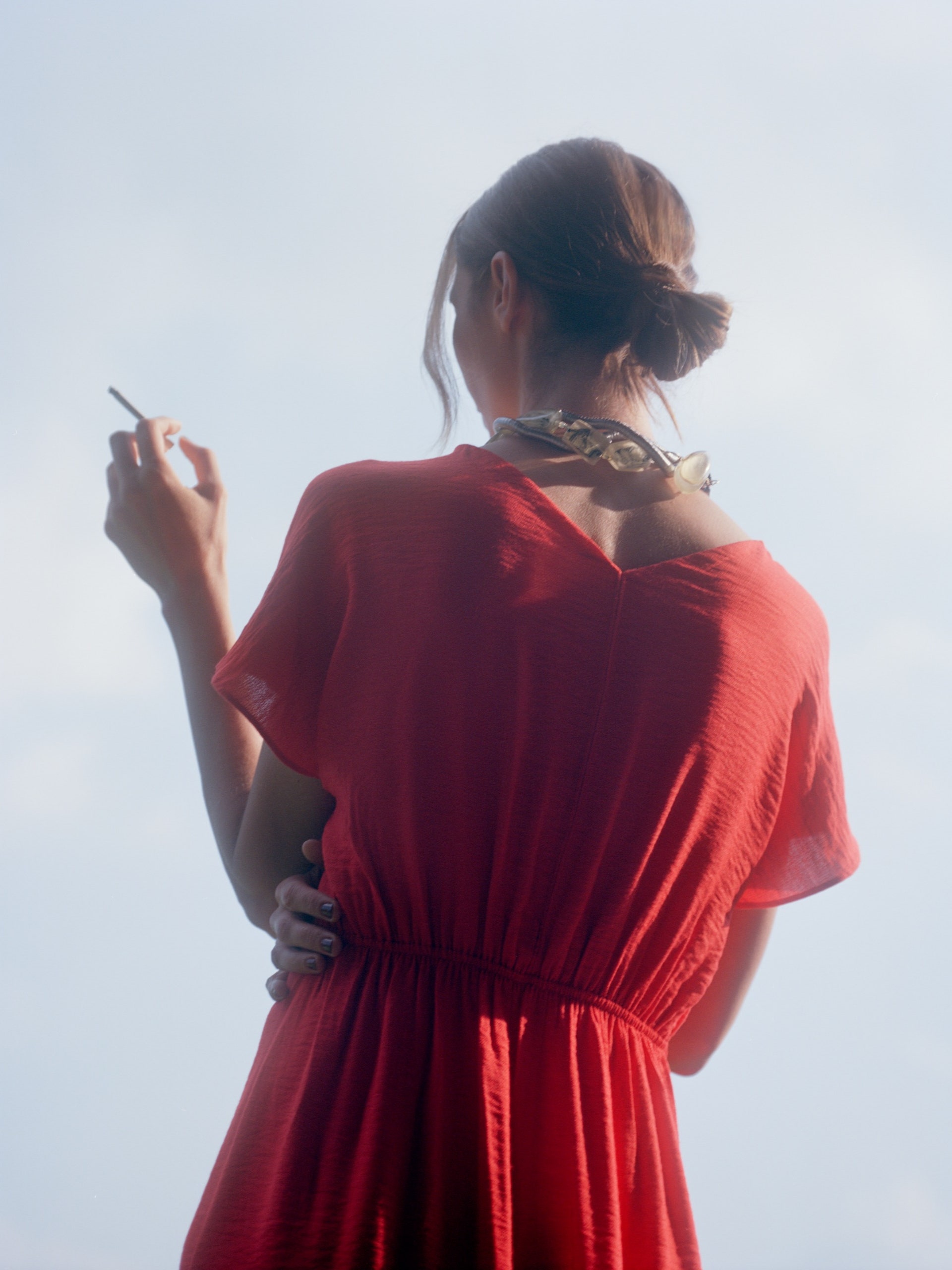 A person wearing an orangered dress with their back to the camera holds a cigarette.