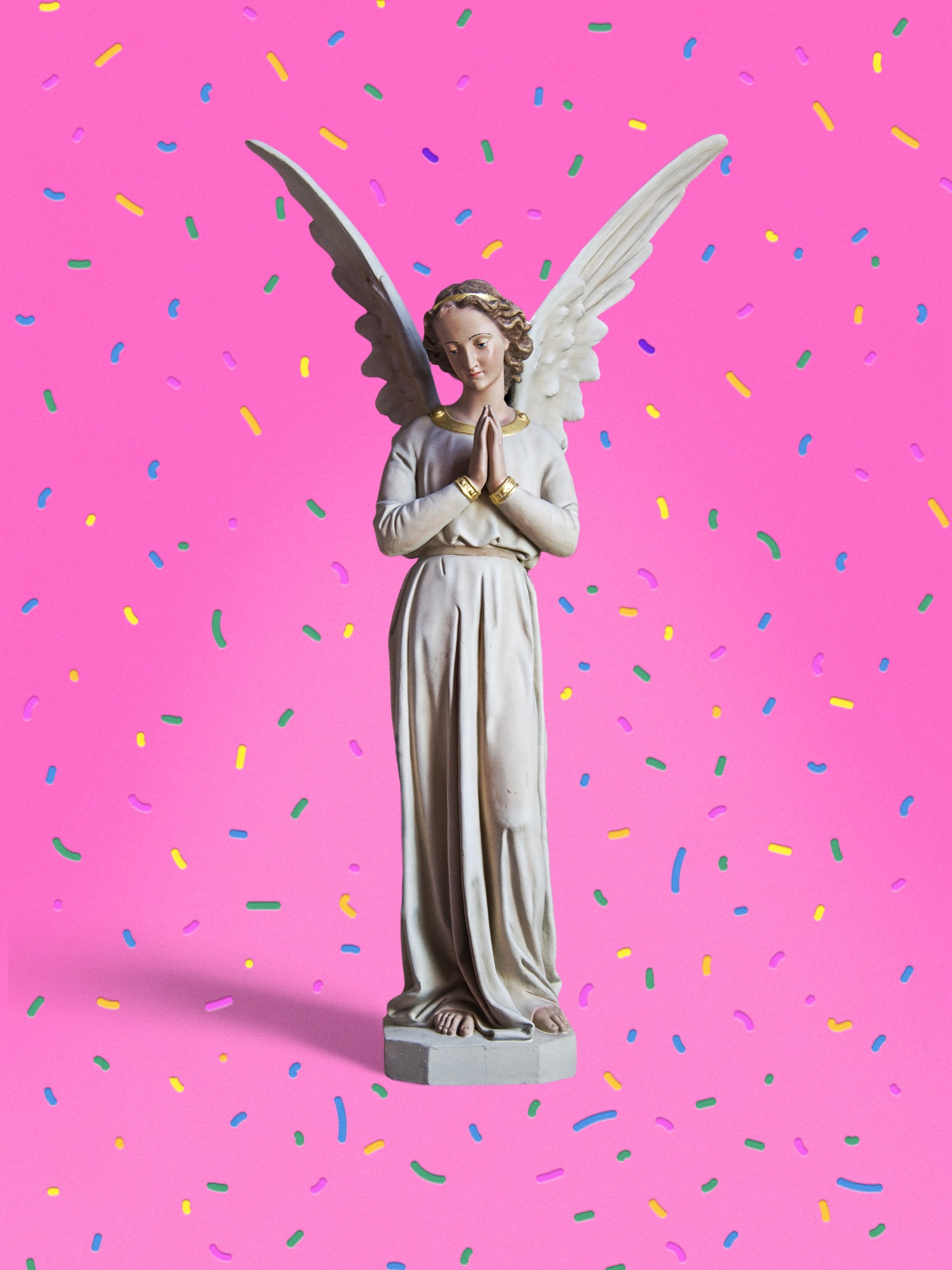 A statue of an angel praying in front of a pink backdrop with confetti.