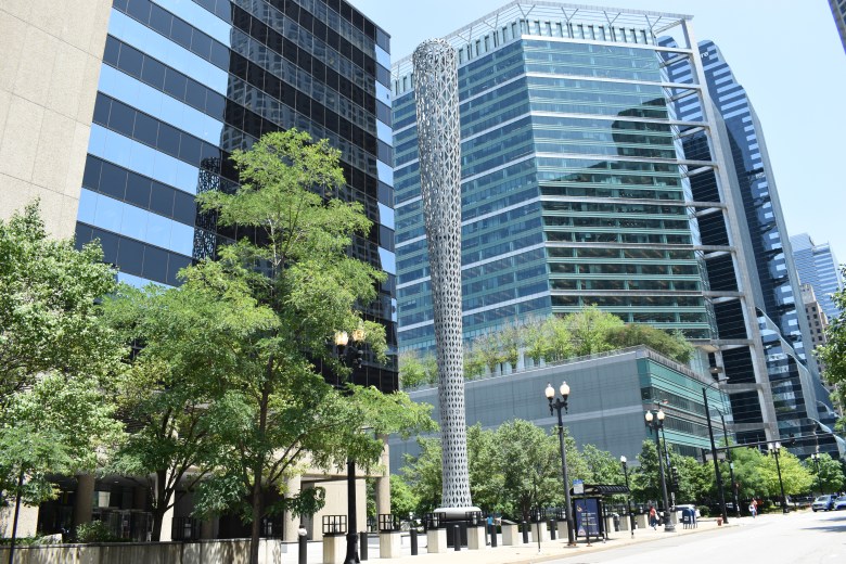 A color photo of Batcolumn, showing glass skyscrapers behind it, and trees on the sidewalk to the left.