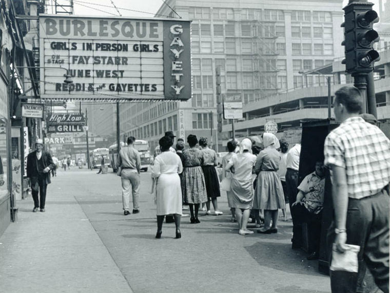 A black and white photo fro, 1960 shows a marquee for Gayety hanging over the sidewalk. Many people are walking or waiting on the sidewalk. Behind the marquee is a sign for High Low Grill.