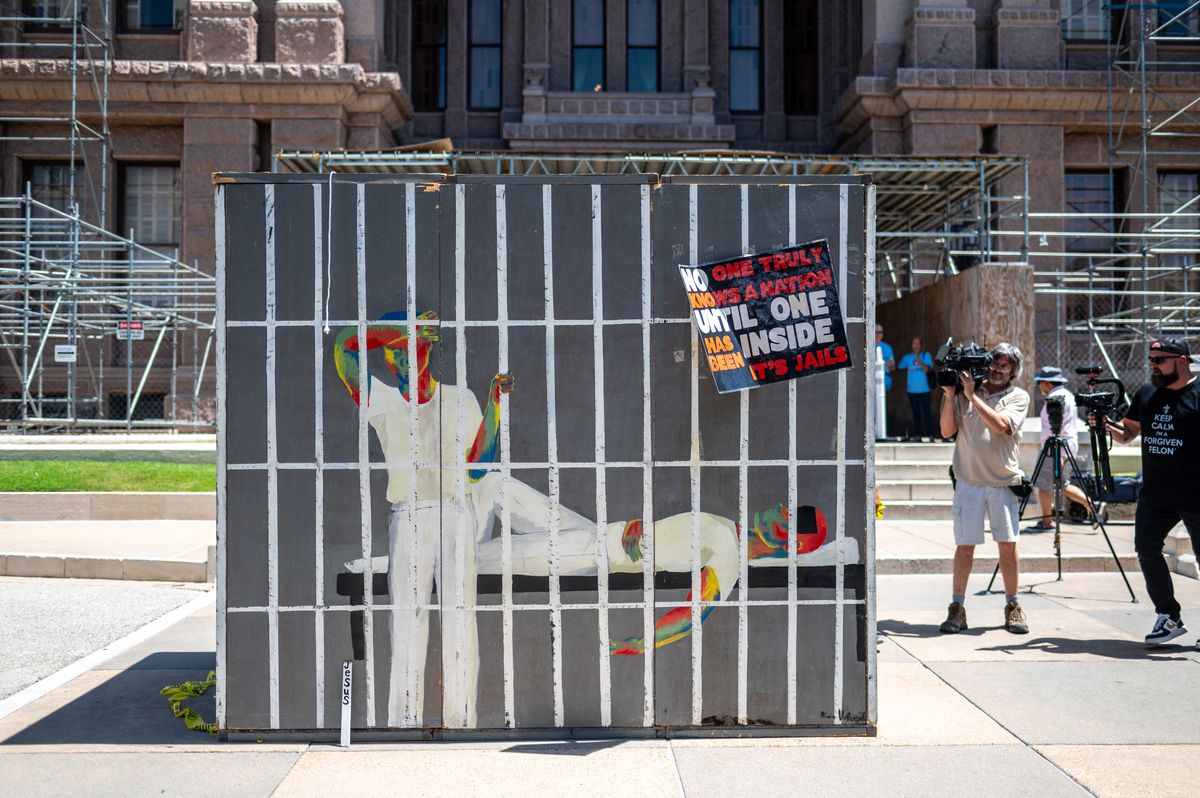 A mock prison cell made of cardboard and paper stands in front of the Texas state Capitol building in Austin, with onlookers and cameras nearby. 