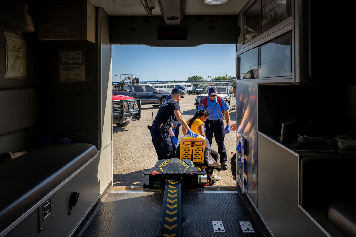 A view from inside the back of the ambulance shows several officials and paramedics lifting a pregnant woman in an emergency chair into the back on a ramp.