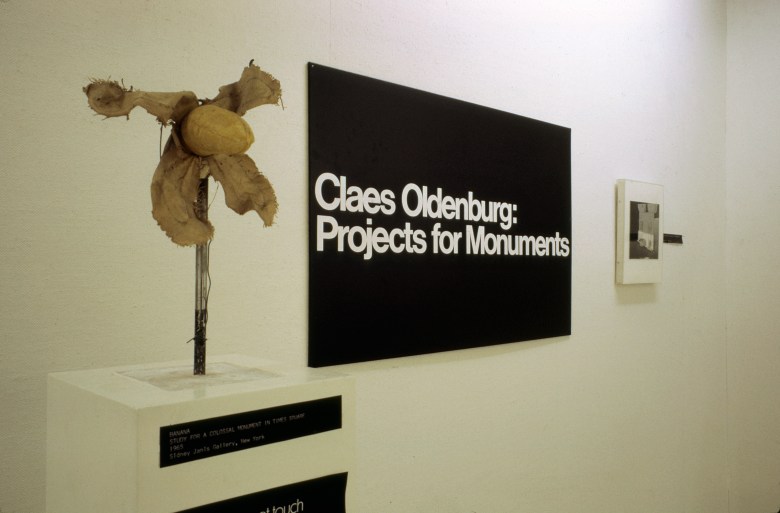 Installation photo has a black sign in the center with the name of the exhibition. To the left is a cloth study of a Times Square monument, resembling an unpeeled banana.