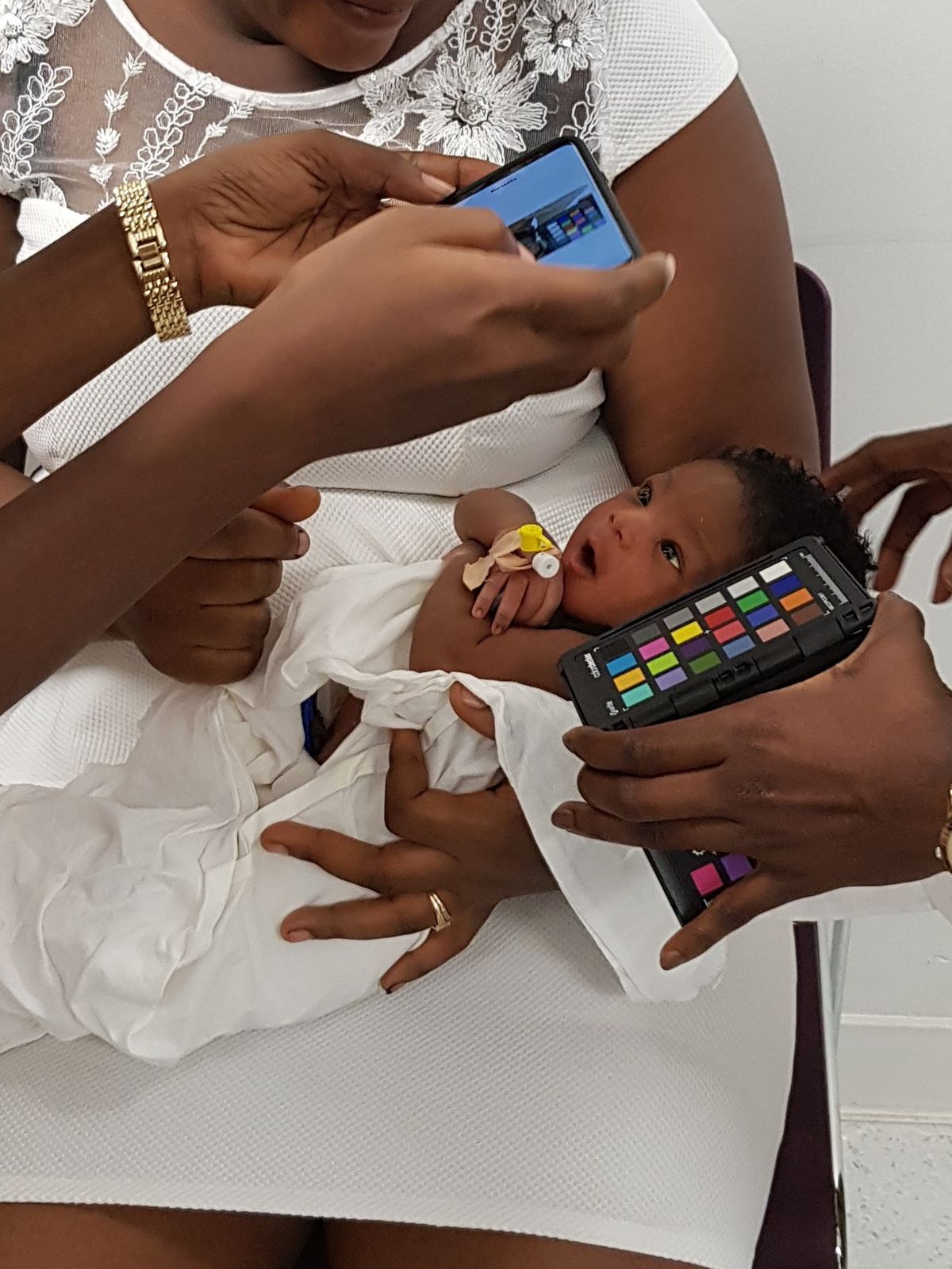 A photo of a mother wearing a white dress holding her baby. One person’s hands hold a color checker card next to the baby’s face while another person’s hands take a picture on a smartphone. 