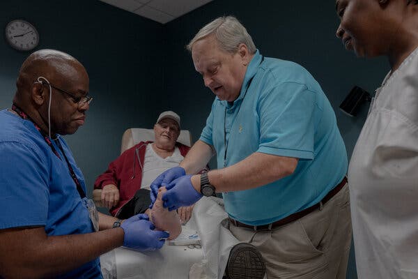 A photograph of a man lying on a hospital chair while three people look at his toeless foot. Dr. Girnys holds his foot with blue gloves, inspecting the area where the big toe once was. 