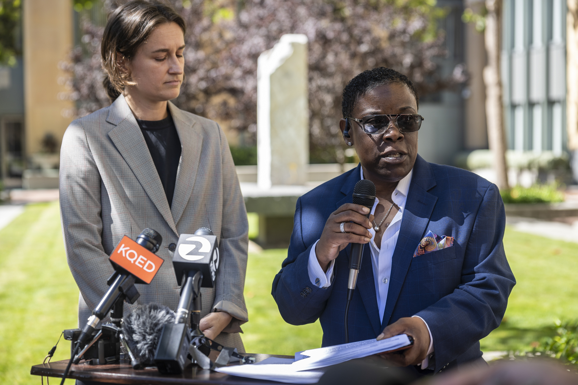 A person in sunglasses speaks into a microphone in an outdoor setting.