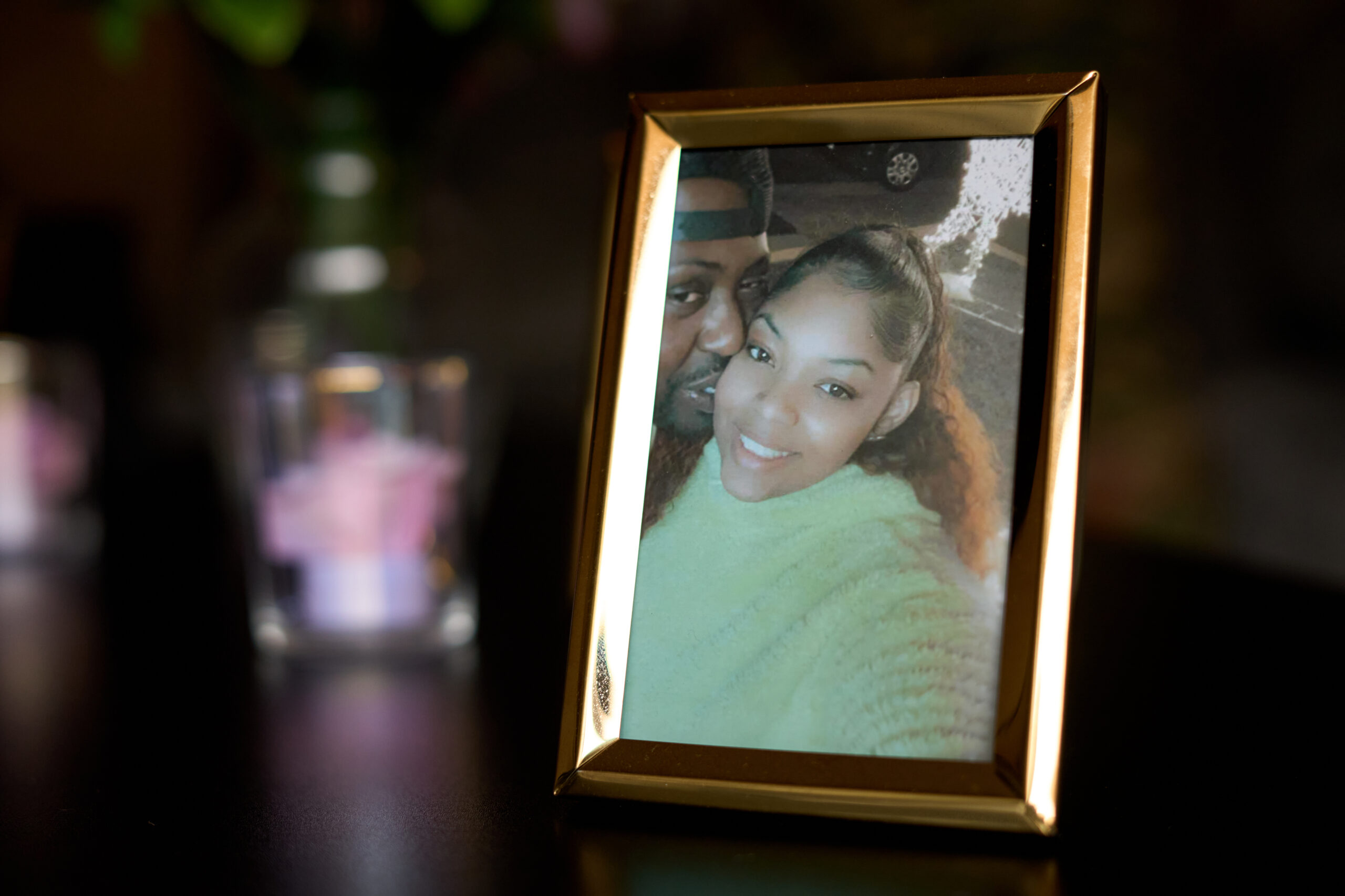 A gold frame on a table shows a photo of a Black couple, a young man with a beard and a young, smiling woman her hair tied back in a ponytail. The man is kissing the woman on the cheek. 