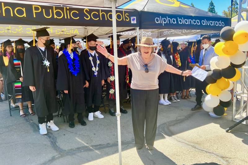 A woman wearing a hat with her arms outstretched stand behind students in a graduation gowns under a tent that reads 