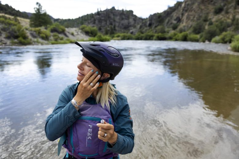 A woman wearing a lifejacket and helmet stands in a river back puts glitter on cheekbones
