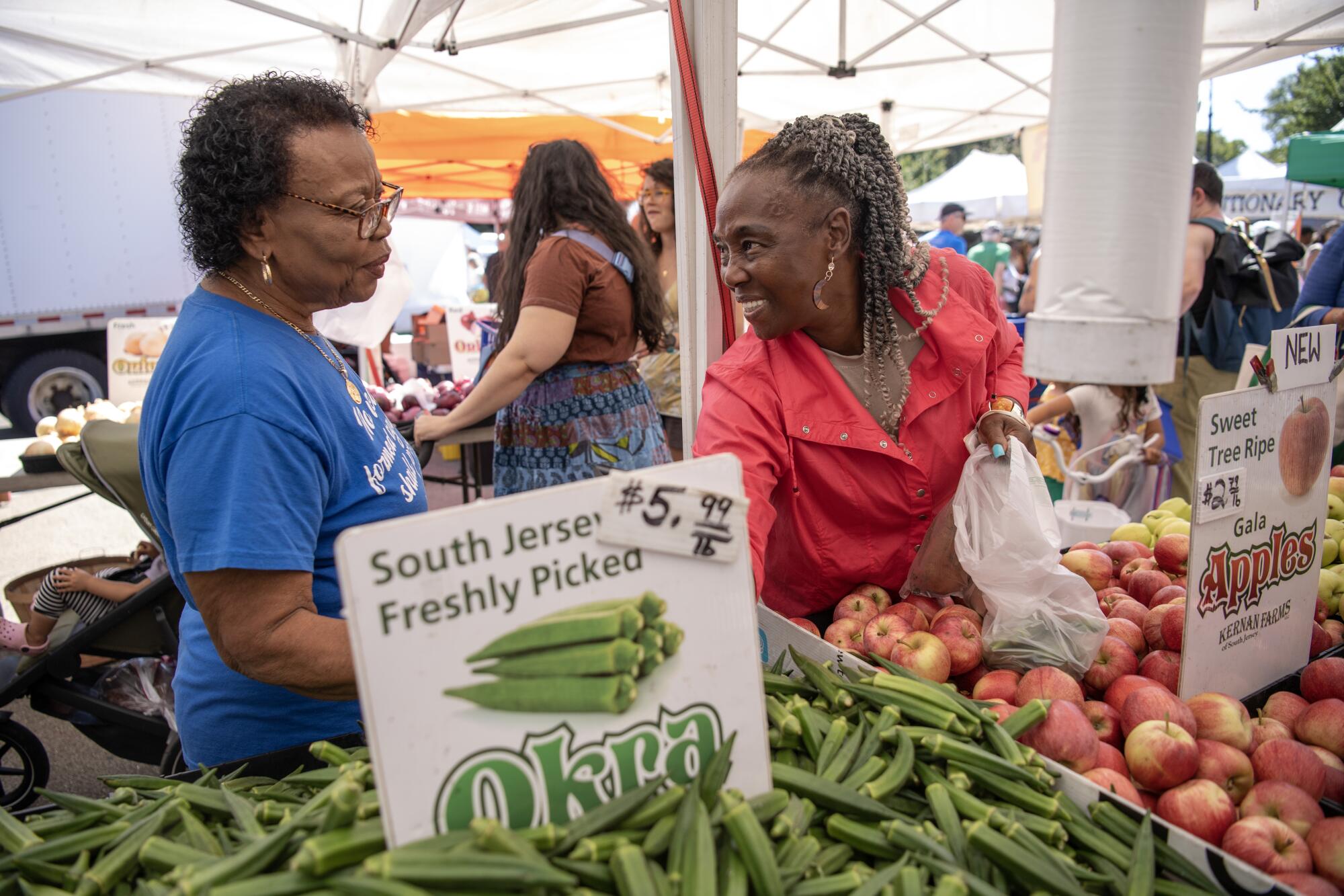 Two women talking at a farmers market, one holding a plastic produce bag and reaching into a pile of okra in the foreground