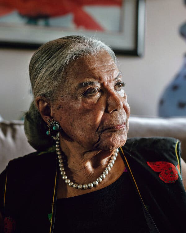 A woman with salt-and-pepper hair, wearing earrings and a strand of beads, poses for a photographer.