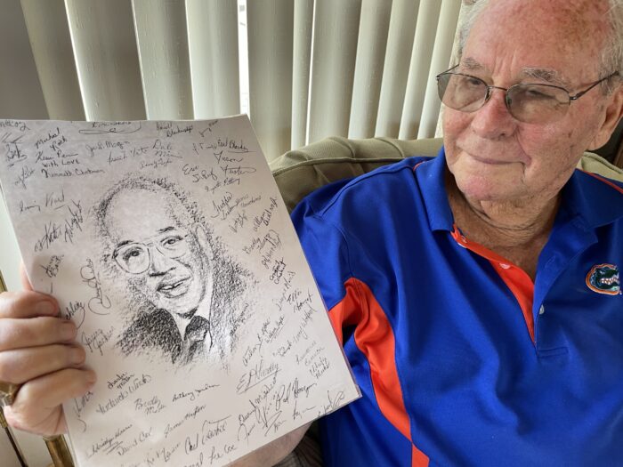 A man in glasses and wearing a blue shirt holds up a drawing of another man. The page has several signatures on it.