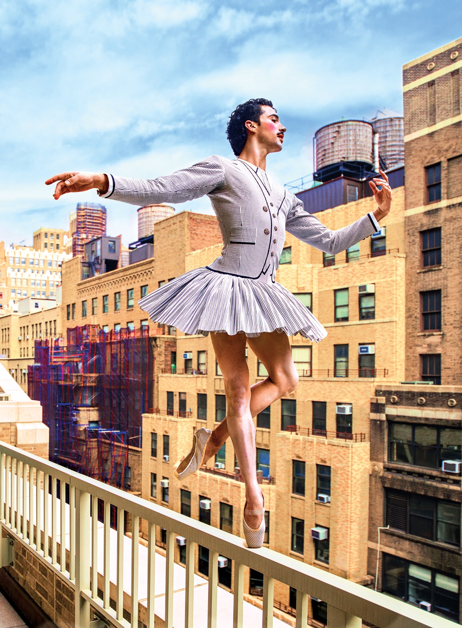 A male ballet dancer wears a lavender sweater and skirt while balancing on the railing of a balcony.