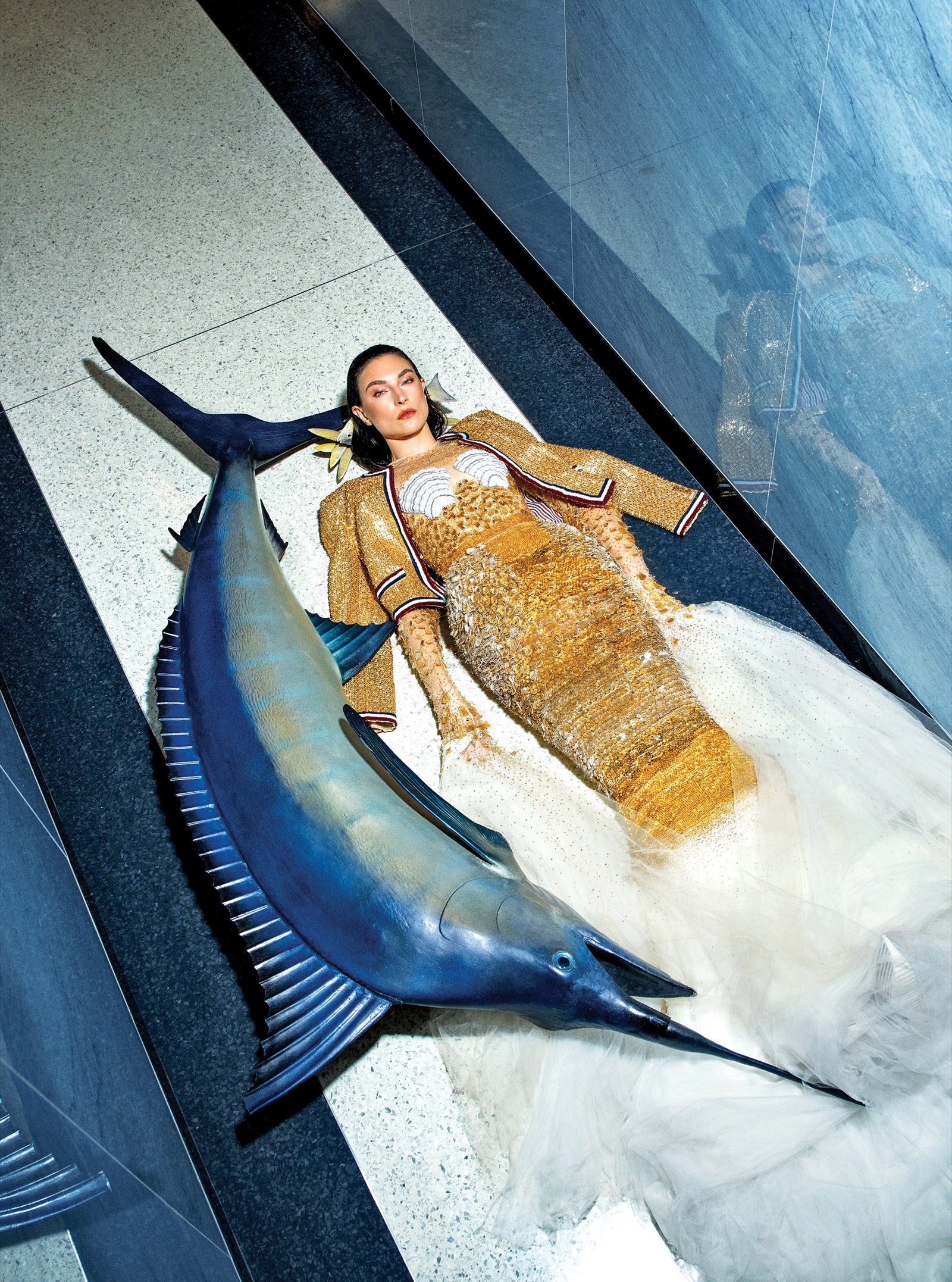 A model lies on the floor in a yellow beaded dress beside a large decorative swordfish.