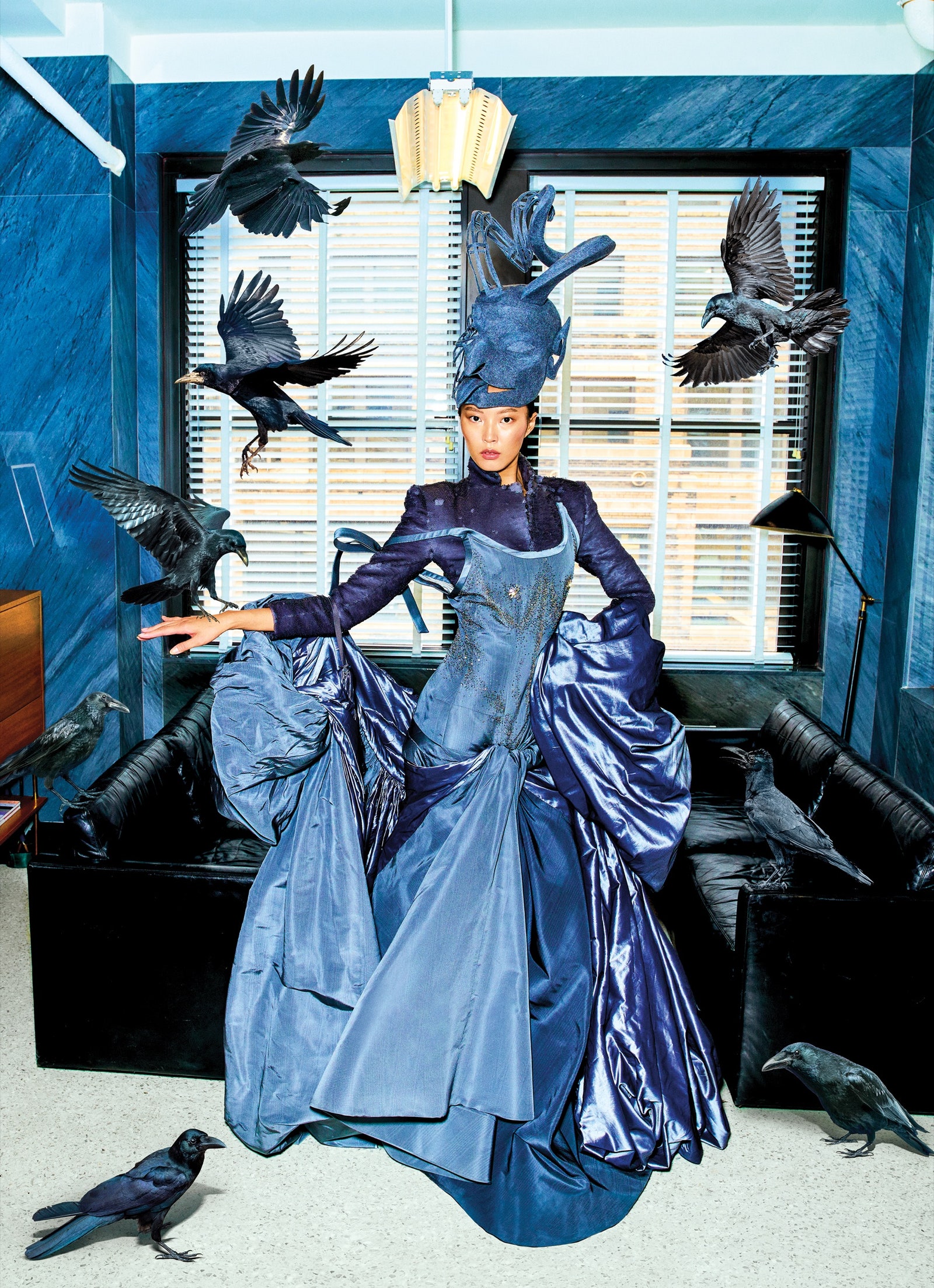 A model poses in an elaborate blue gown while birds fly and perch around her.