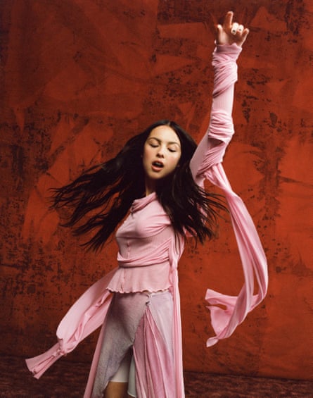 Olivia Rodrigo standing against a dark red background with her arm in the air, wearing pink dance wear and shoes