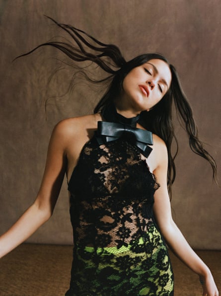Olivia Rodrigo with her eyes closed, her head on one side and her hair in the air, wearing a black lace dress with green luminous tights underneath