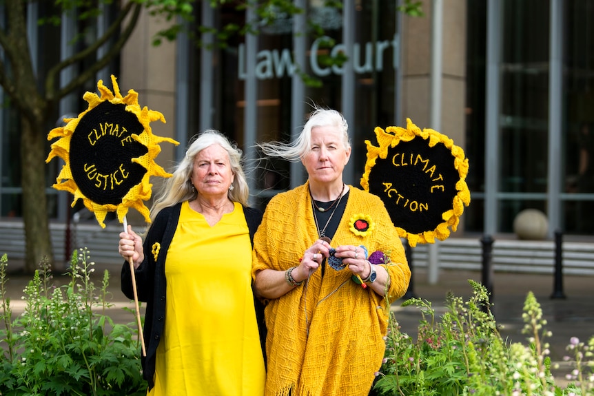 Two older women with grey hair, wear bright yellow clothing and hold signs in the shape of sunflowers reading 'climate justice'