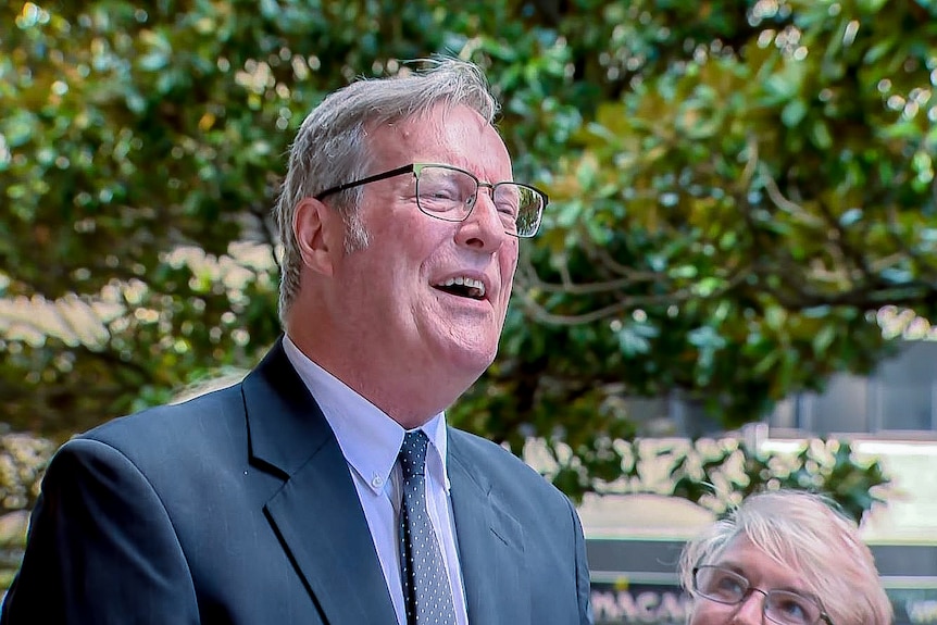 A man with grey hair and glasses, wearing a dark suit, smiles as he speaks to the media. 