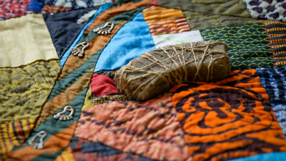 A rock wrapped in twine sits atop a colorful patchwork quilt, with silver footprint beads attached in a pathway just to the side.