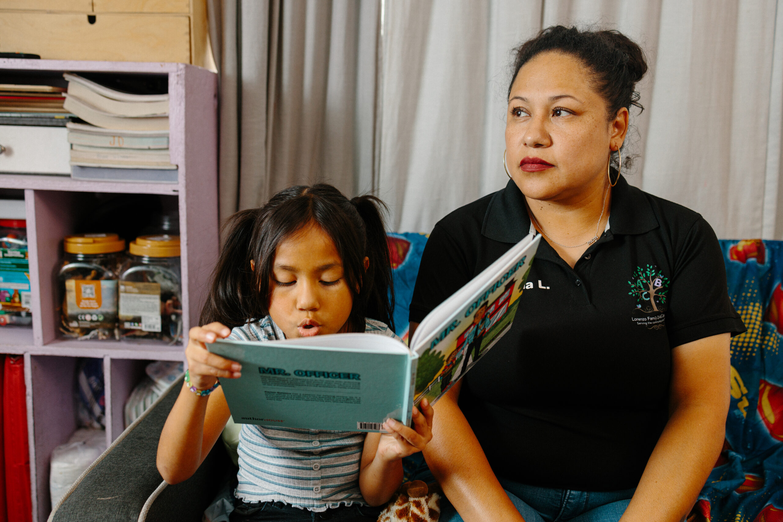 Adriana Lorenzo poses for a portrait at her home based daycare, The Lorenzo Family Child Care while her daughter reads a book next to her.