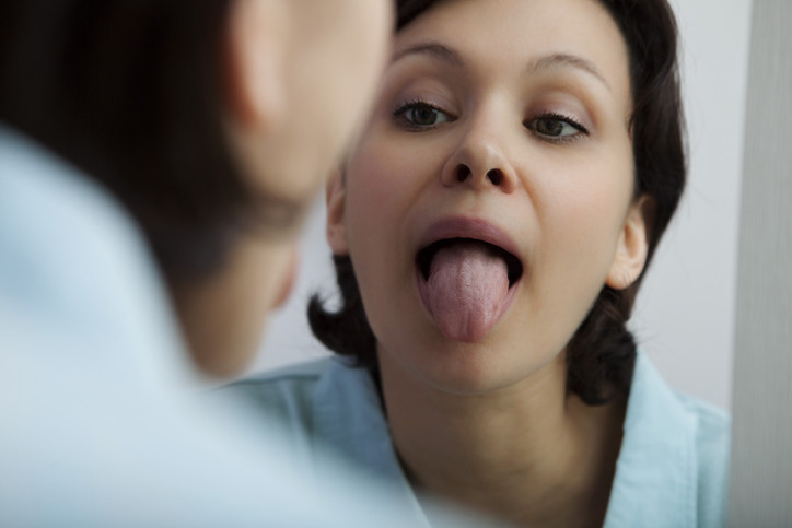 A woman with brown hair, brown eyes, and a blue shirt is reflected in a mirrow as she sticks out her tongue