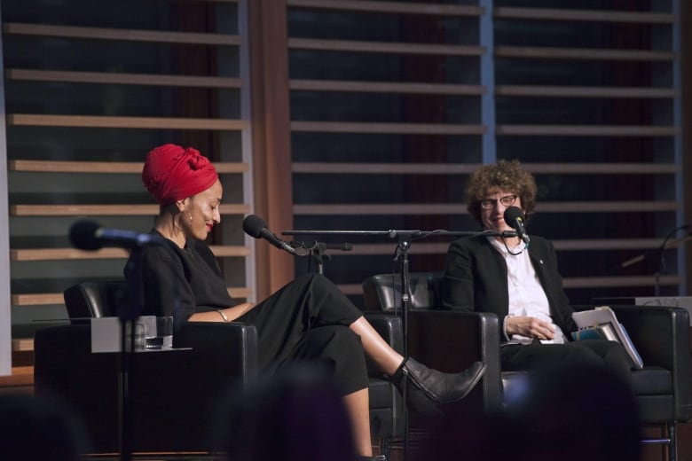 Two women sit on chairs on a stage, the one on the left wears a red headscarf, the other has short curly brown hair. 