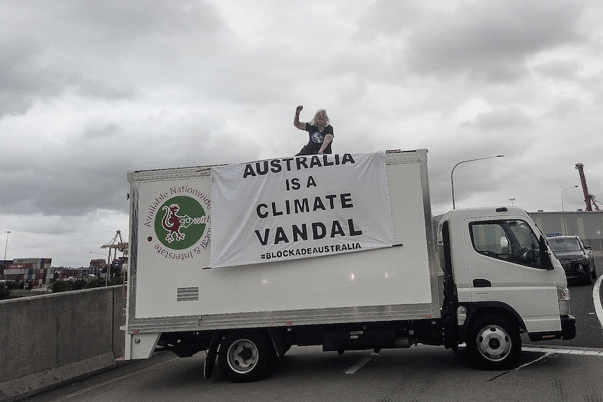 A woman with white hair sits on top of a white truck, raising her fist, a sign saying 'Australia is a Climate Vandal' hangs.