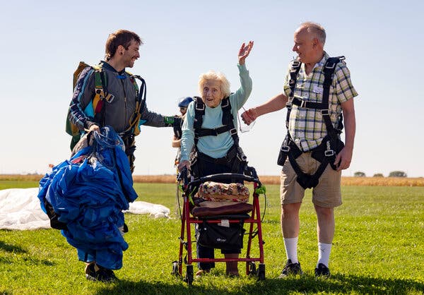 Dorothy Hoffner, wearing a baby blue sweater, holds on to a walker as she waves from the field where she had landed. Her tandem jumper, Derek Baxter, and a friend, Joe Conant, stand on each side.