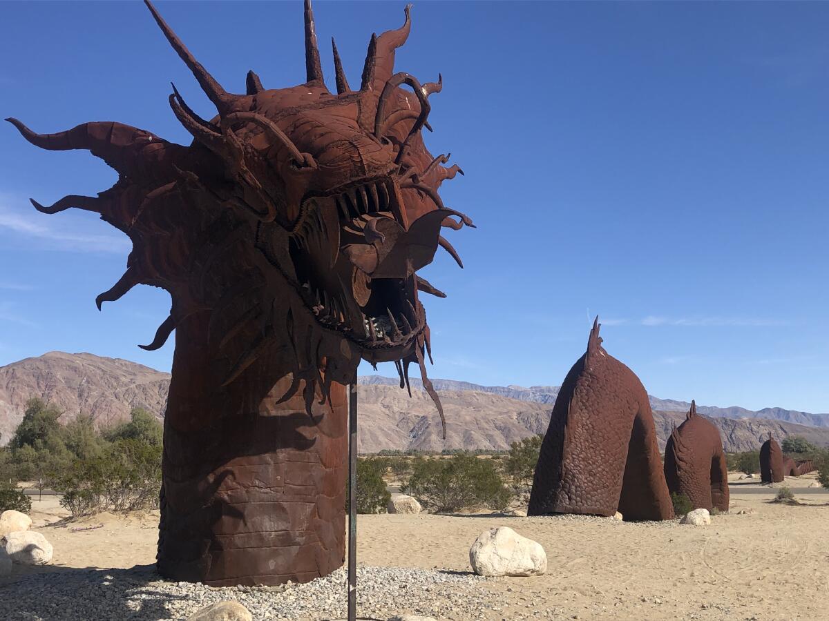 A brown-colored sculpture of a dragon's head and body rising from a sandy ground, with mountains in the background 