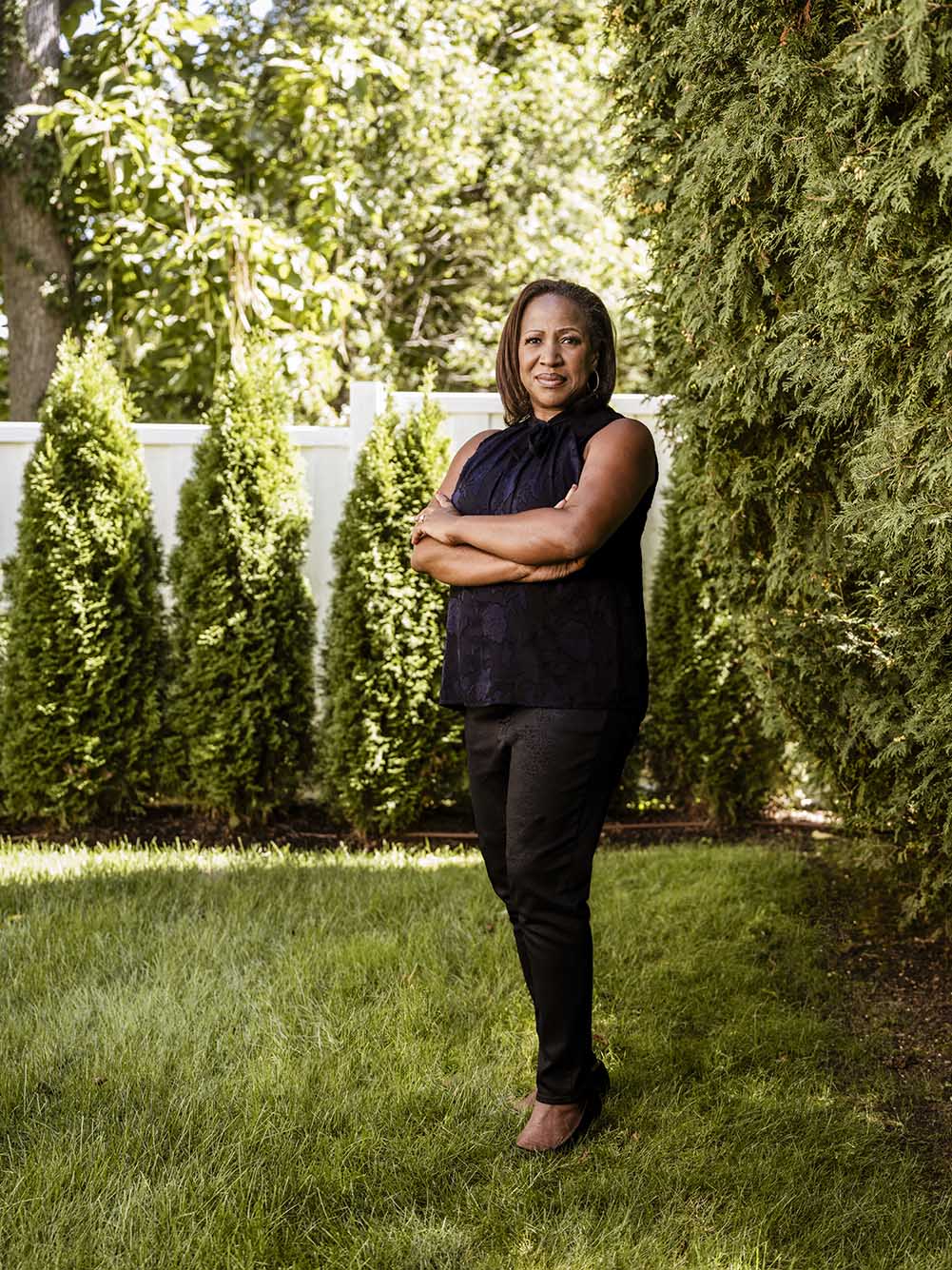 Photo: Charlene Coyne, a Black woman wearing a black blouse and black pants, smiles and poses with arms crossed on a lush, green lawn on a sunny day. Behind her, green shrubs line a tall, white fence.