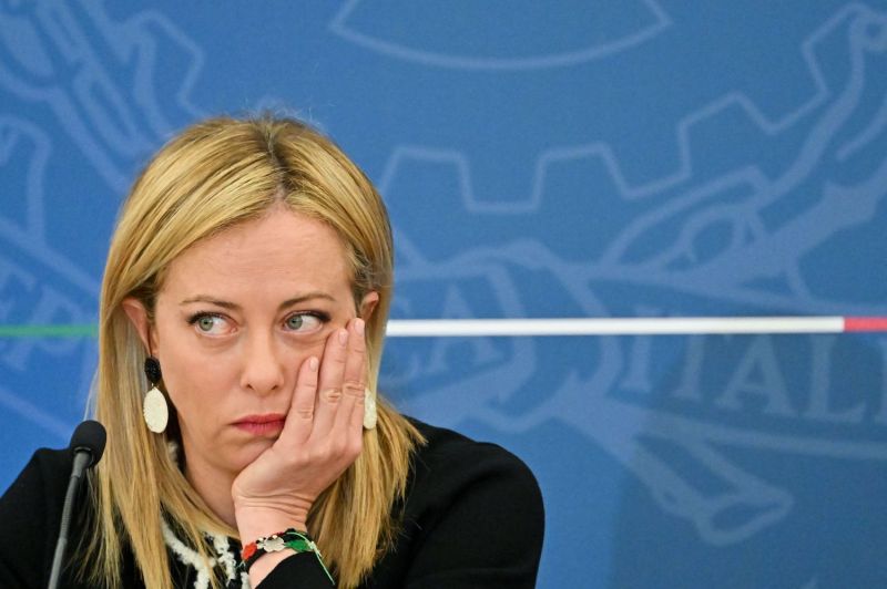 Italian Prime Minister Giorgia Meloni rests her chin on her hand as she sits in front of a microphone during a press conference in Rome. Meloni, a woman in her 40s wearing a black suit jacket, frowns slightly as she looks to the side.