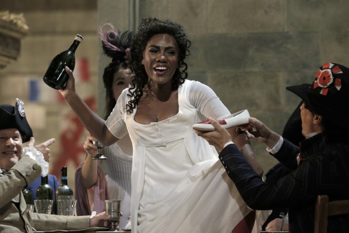 A vivacious black woman in a white dress holds a bottle of liquor as she passes clapping men who are dressed in Napoleonic-era costumes 