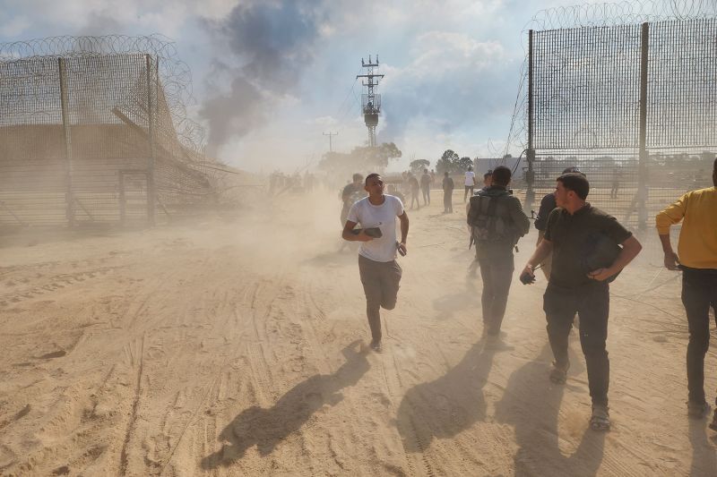 Palestinians run across a dusty landscape through a breach in a high wire fence topped with barbed wire on the Israel-Gaza border.