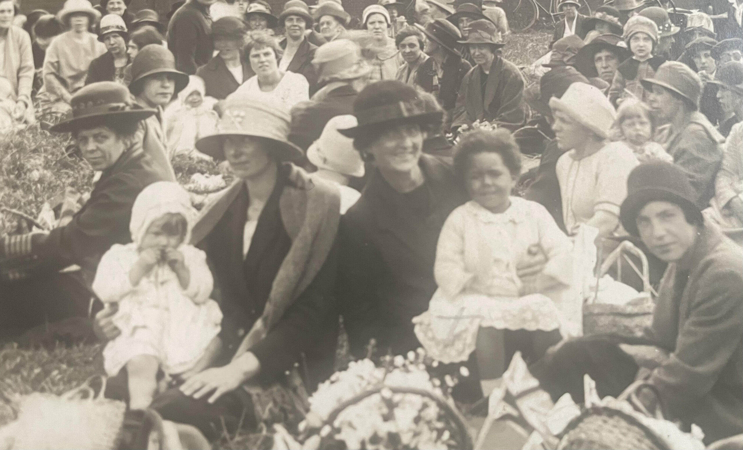 A black and white image of people sat in a park. In the centre, a Black toddler sits on the lap of a white woman in a hat