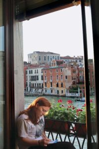 A student sits in the window of Casa Artom studying. Behind her is a window box with red flowers and the Grand Canal and Venician buildings.