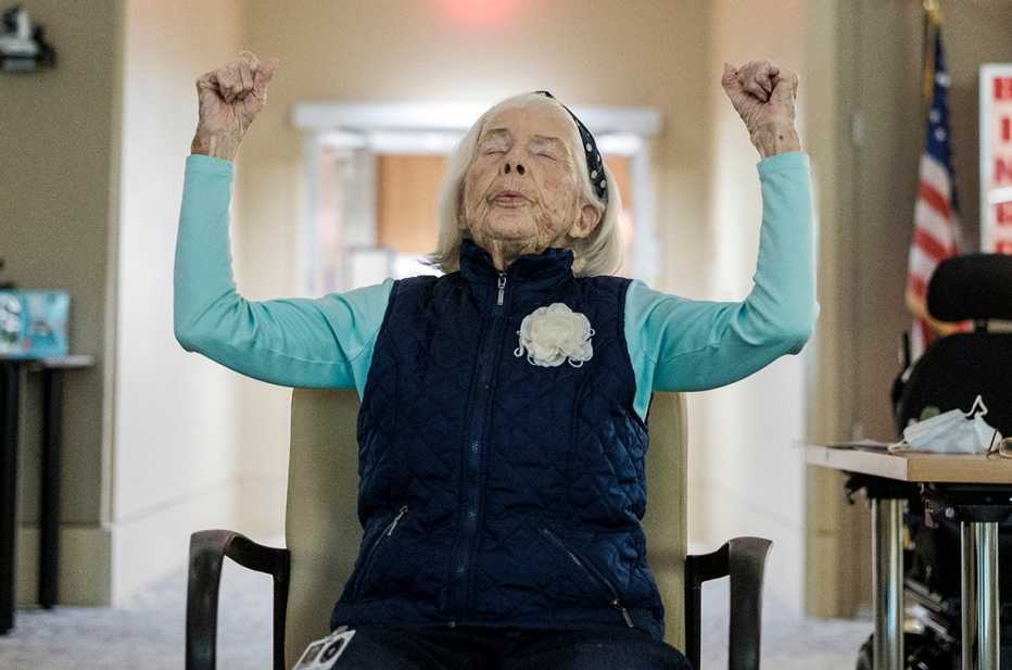 emmy lu daly age one hundred participates in a meditation class seated in a chair with her arms raised and eyes closed