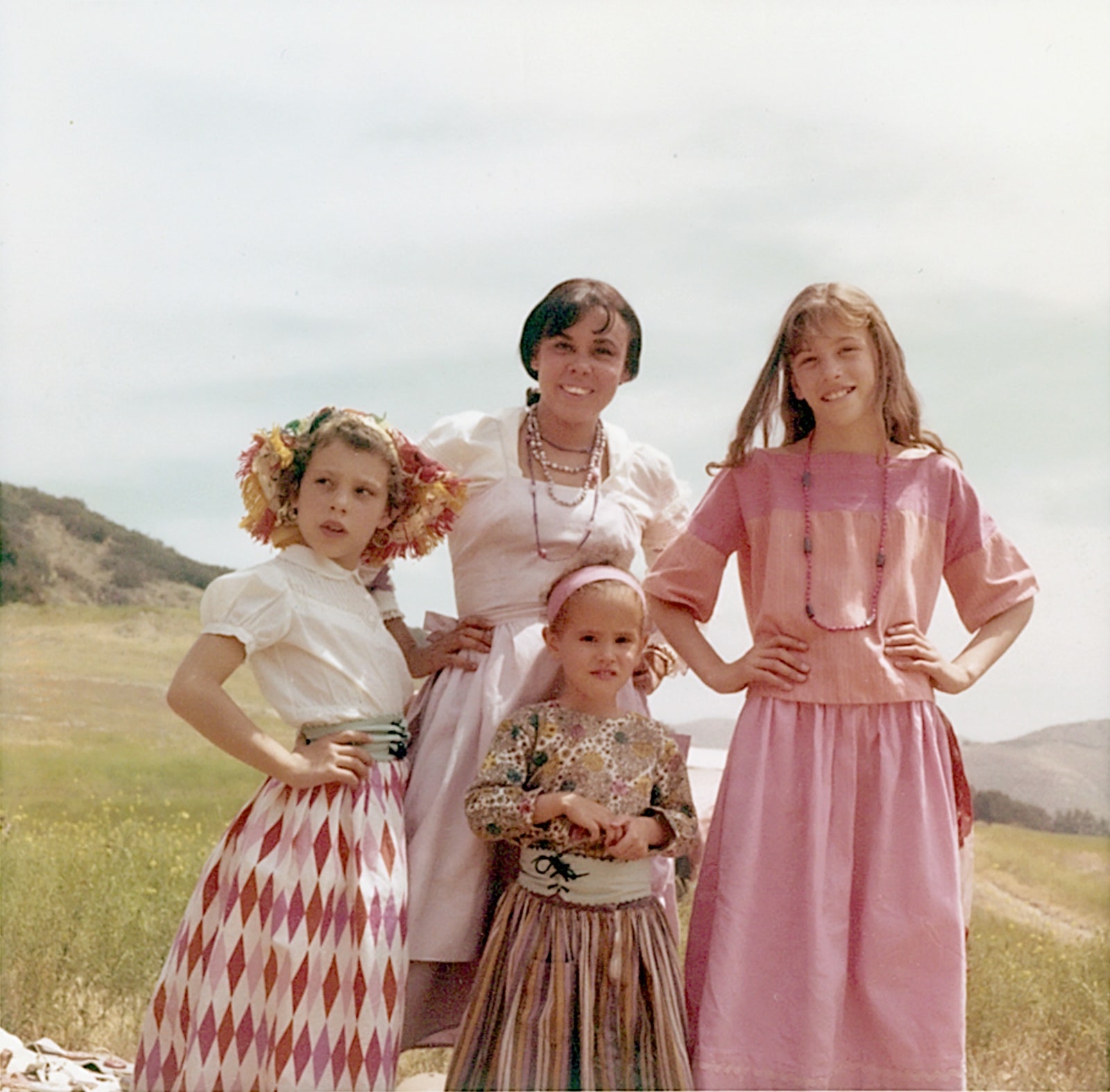 A photograph of a woman with three children standing in a meadow. They are wearing pink and white dresses and three have...