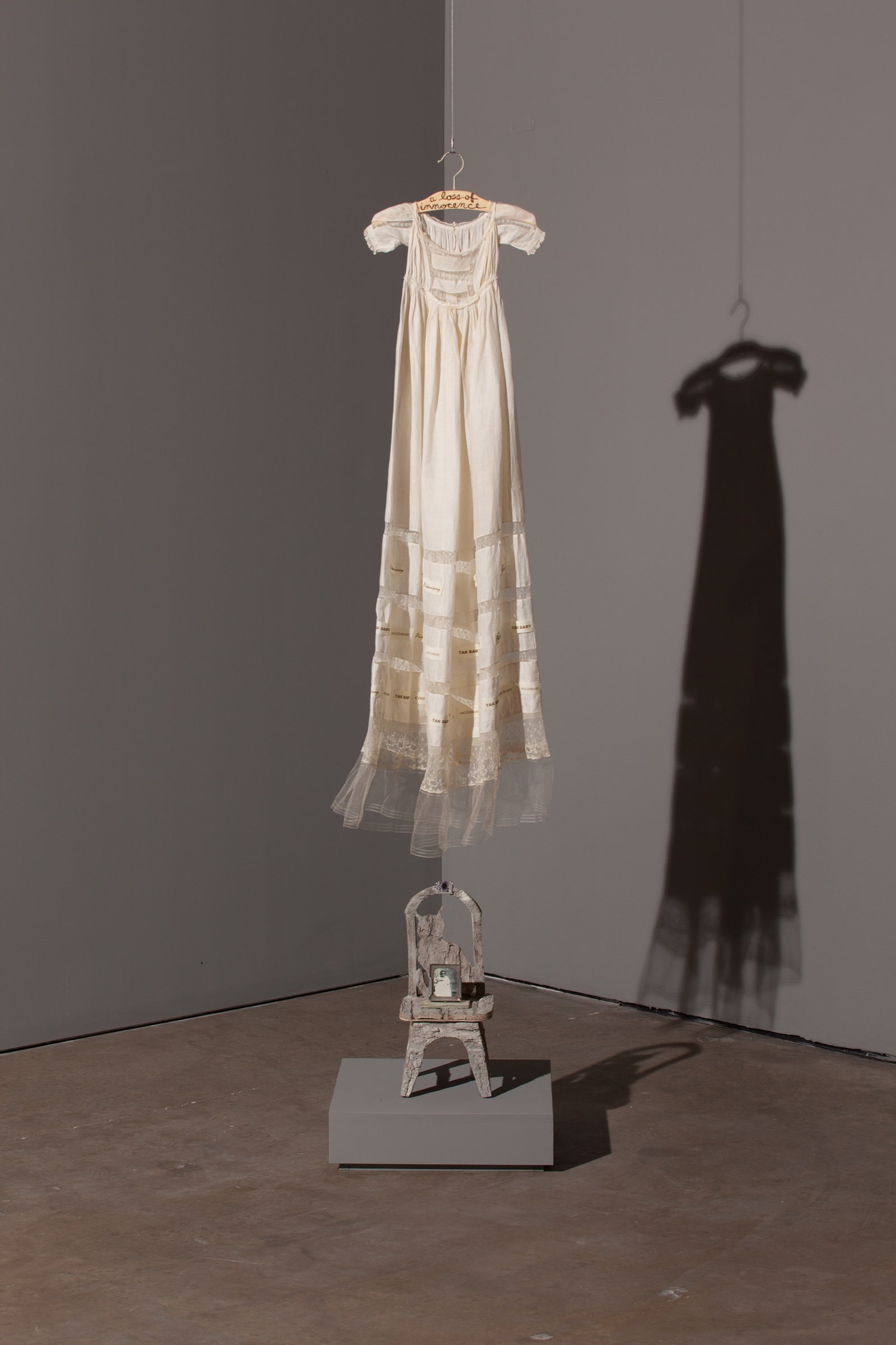 A photograph of an art piece by Betye Saar. The picture depicts a white dress on a hanger above a miniature chair...