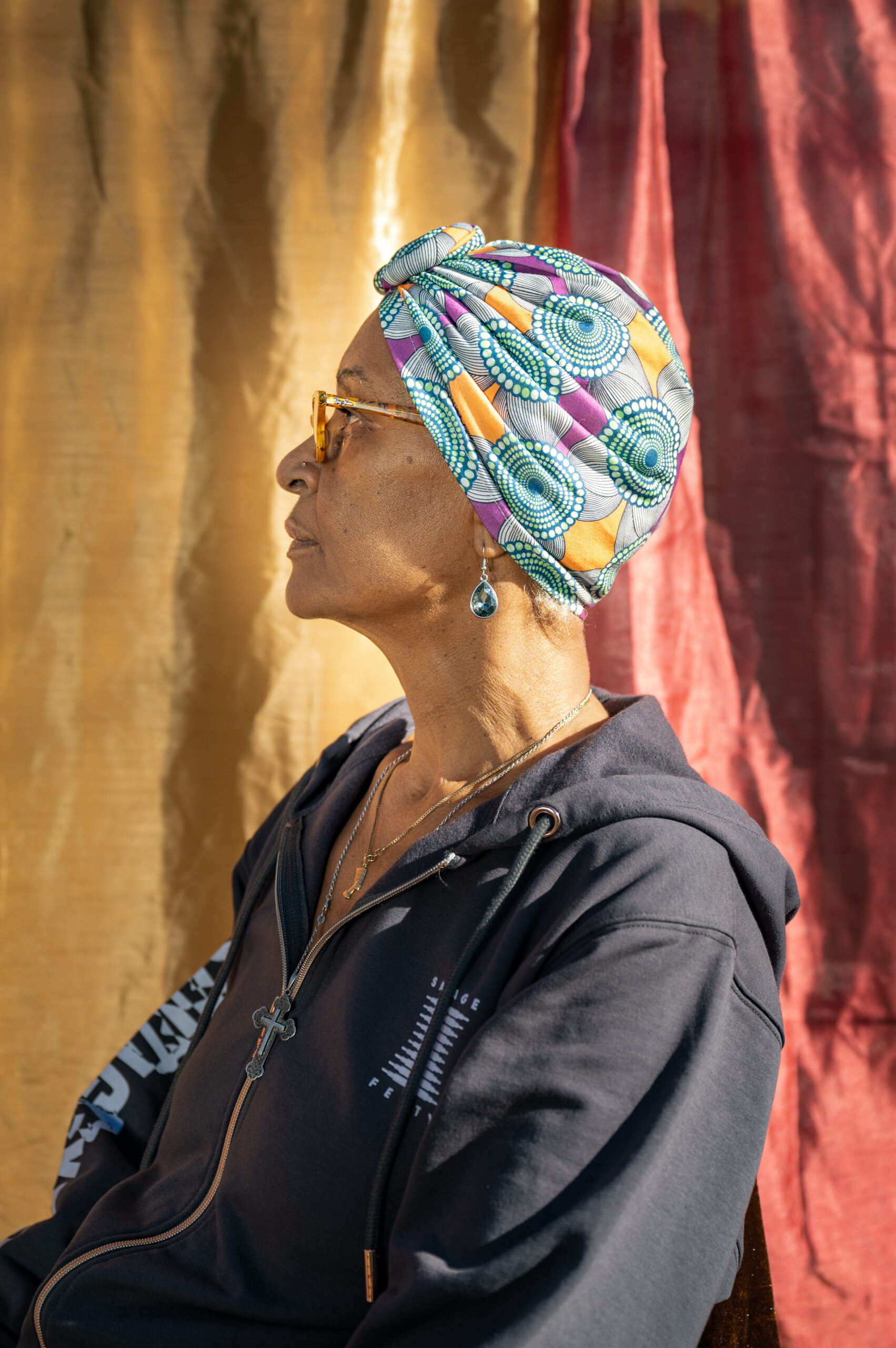 An older Black woman sits in profile, wearing a blue hoodie, gold metal glasses and a carefully tied blue, yellow white and purple patterned scarf tied over her gray locks. An earring with a blue tear-drop shaped jewel is visible in her left ear.