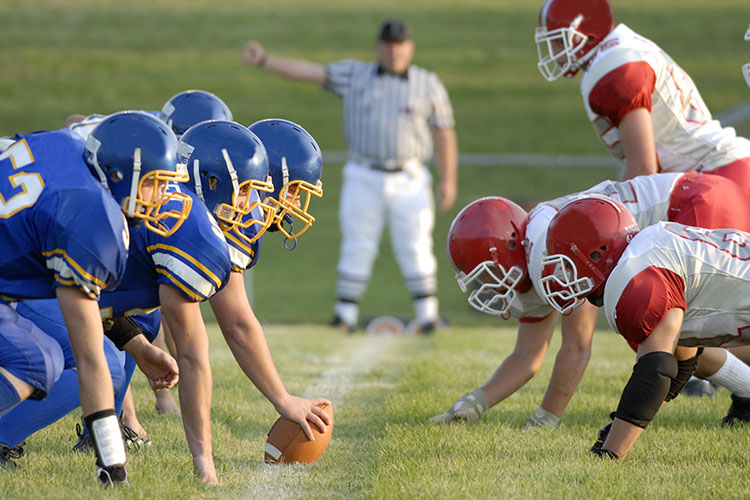 Two football teams face each other, with the blue team touching the football, and the red team on defense. An umpire is visible in the blurred background.