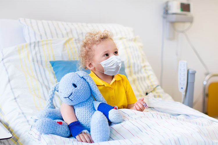 A boy wearing a mask with his arm around a blue stuffed bunny looks offscreen while sitting in a hospital bed.