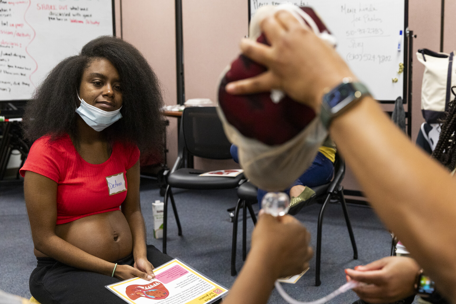 A pregnant African American woman looks on as someone handles a catheter in a room with chairs.