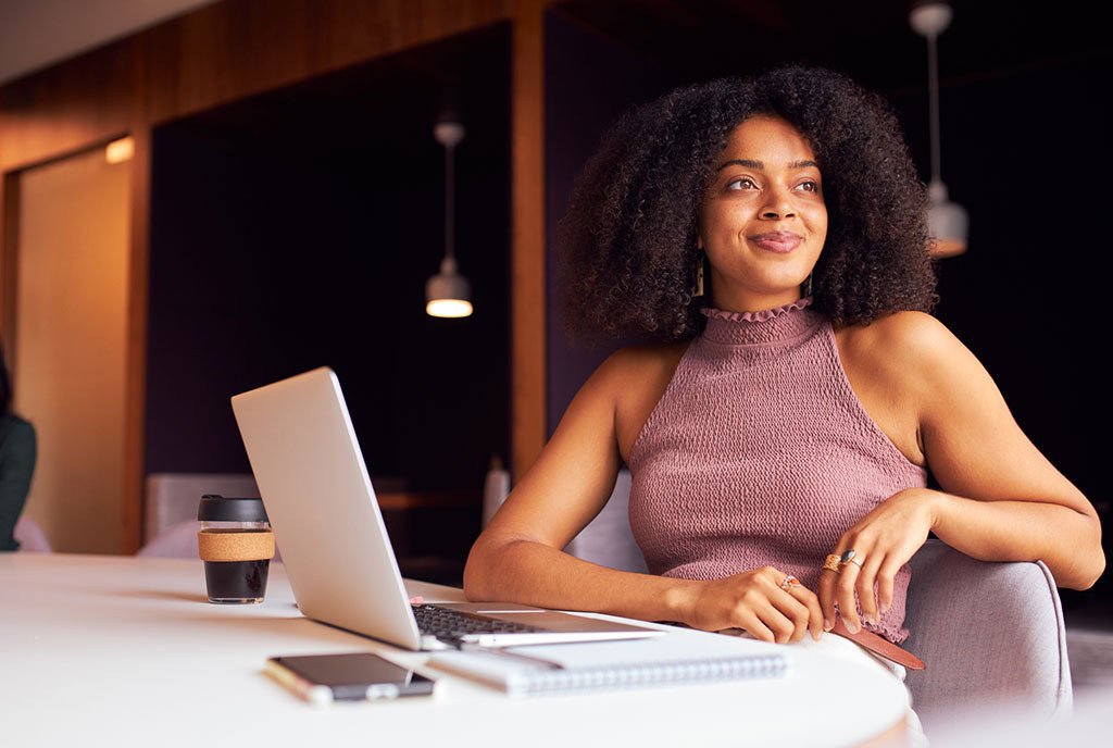 A Black business woman with natural hair, sitting in front of her laptop and looking into the distance with a smile