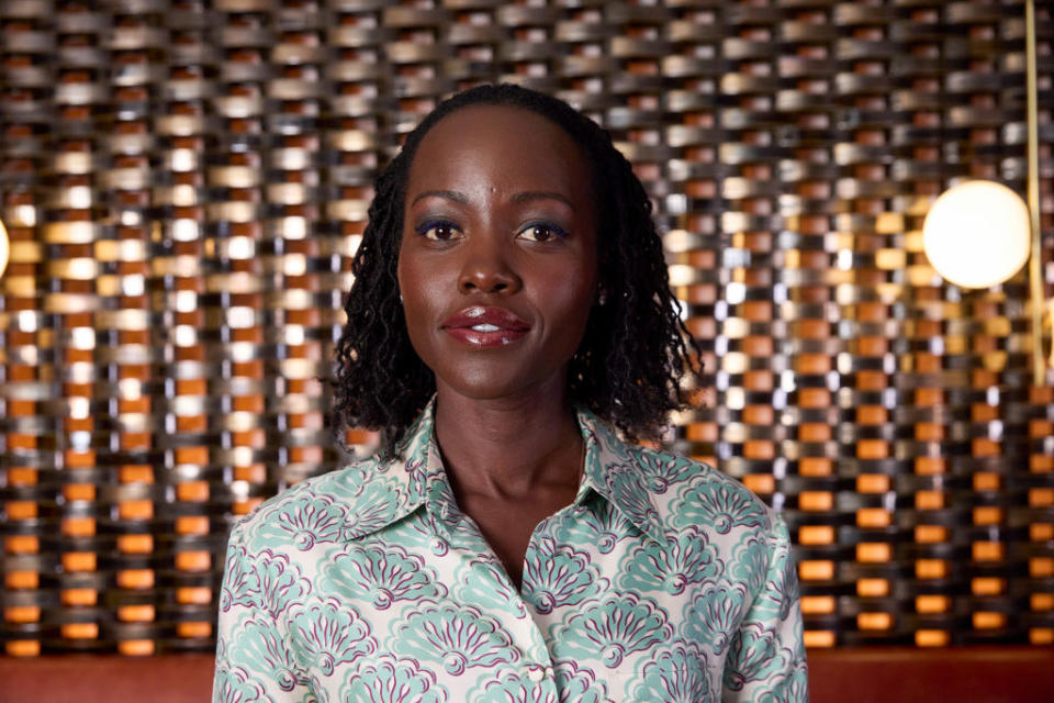 Lupita Nyong'o stands in front of a patterned background, wearing a light-patterned blouse and smiling slightly