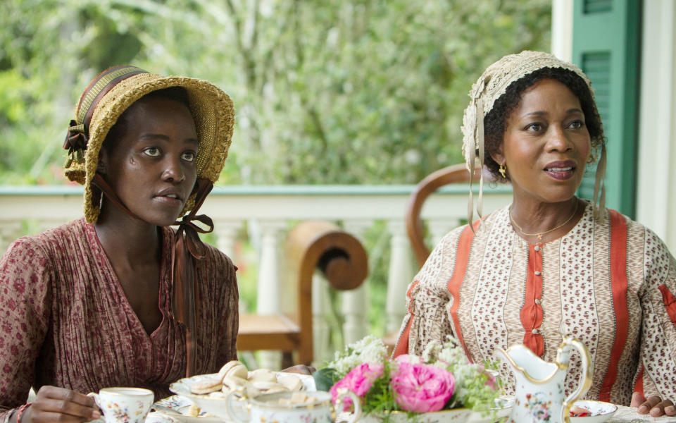 Lupita Nyong'o and Alfre Woodard in historical costumes, wearing bonnets, sit at a table with tea and flowers in an outdoor setting