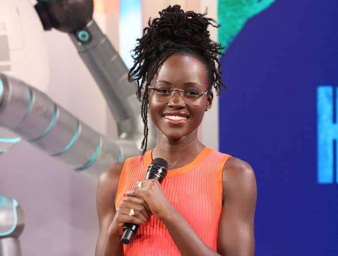 Lupita Nyong’o smiles while holding a microphone. She wears a sleeveless dress and glasses, with her hair styled in an updo