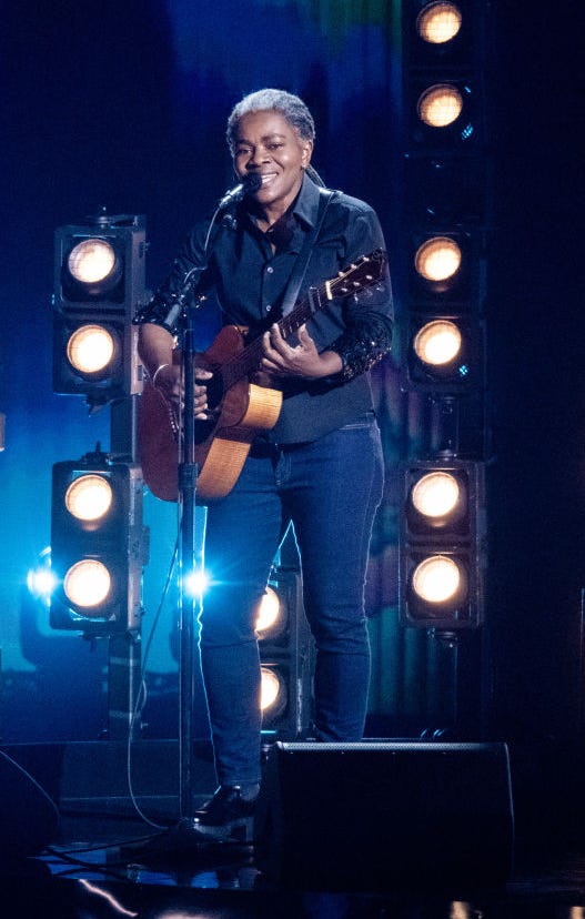 tracy chapman playing guitar and singing in front of yeloow stage lights at the grammy awards